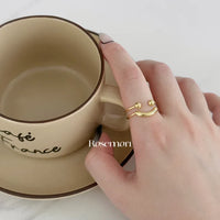 Hand mit goldfarbenem Smiley-Ring hält beige Tasse, stilvoller Schmuck, minimalistisches Design.