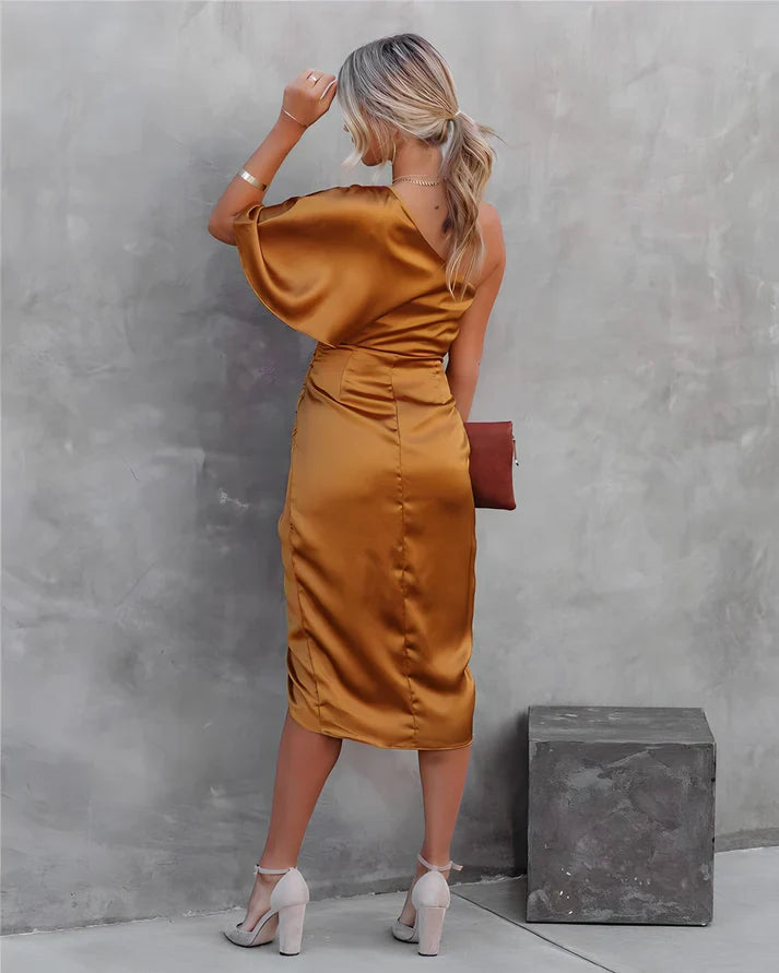 Frau in goldfarbenem Satin-Kleid mit asymmetrischem Schnitt, steht vor grauer Wand.