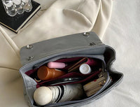 Graue Handtasche mit Kosmetik, Geldbörse und Notizbuch auf beigem Stoff.