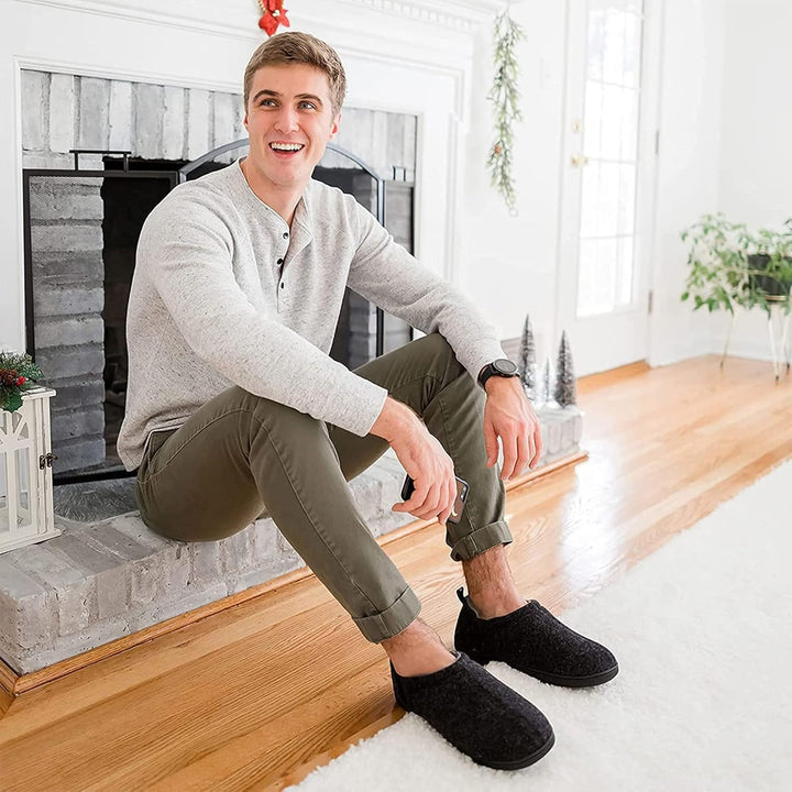 Mann in grauem Pullover und schwarzen Hausschuhen sitzt lächelnd vor Kamin im Wohnzimmer.