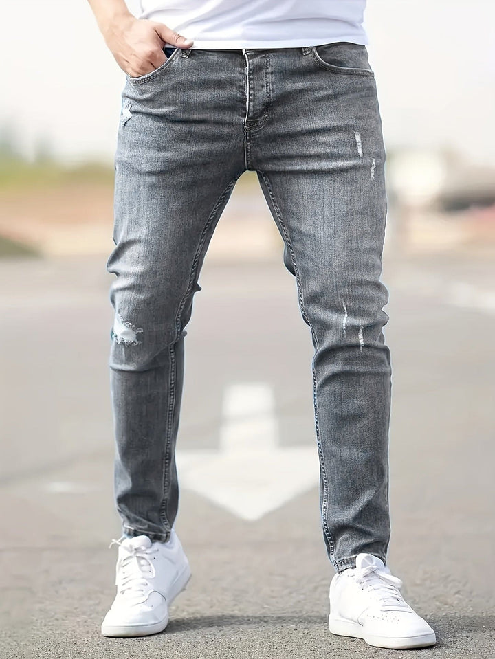 Graue Herren-Jeans, Slim Fit, mit leichten Rissen, kombiniert mit weißen Sneakers auf Straße.