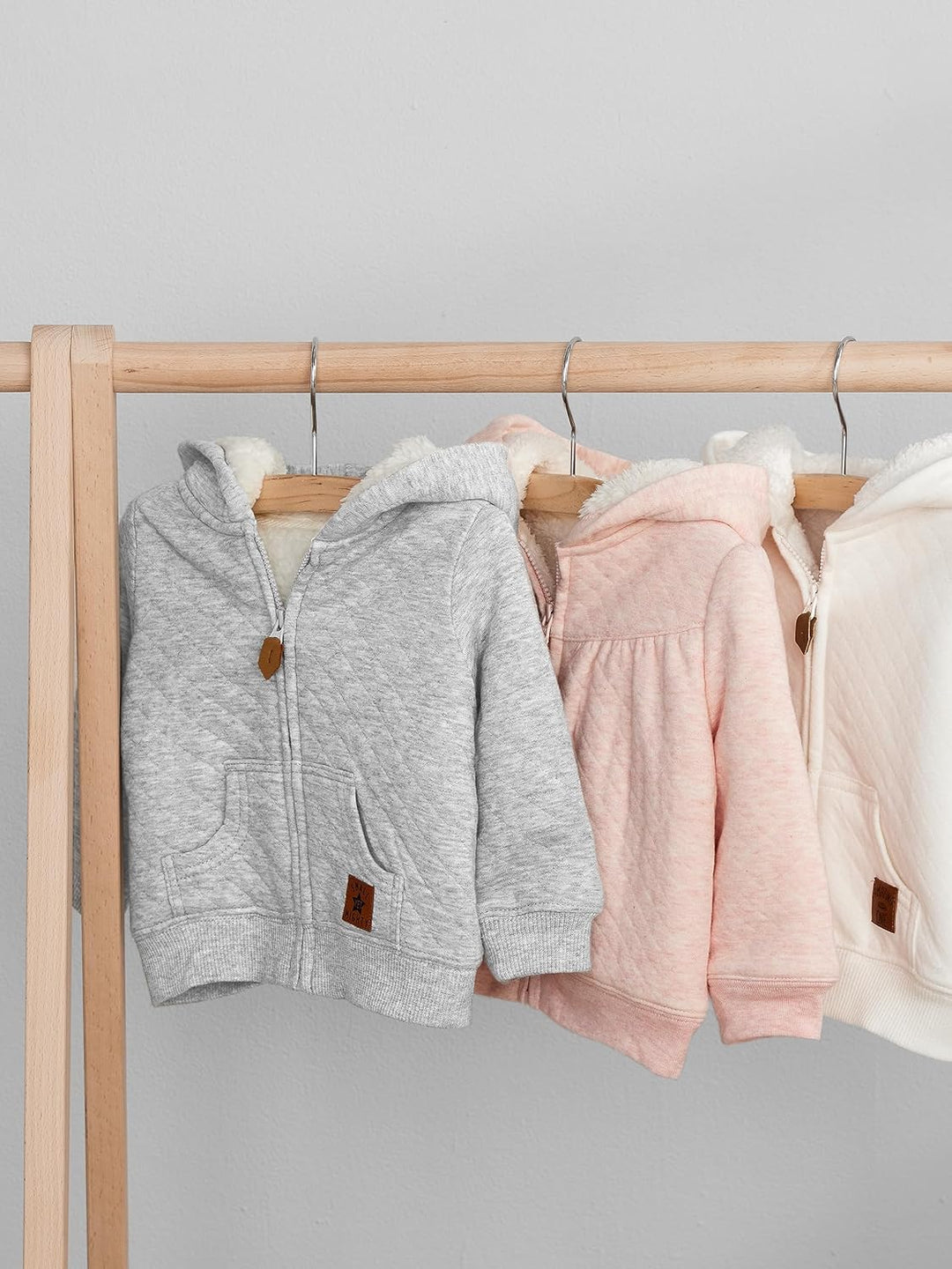 Drei Baby-Kapuzenjacken aus Baumwolle in Grau, Rosa und Weiß auf Holzständer.