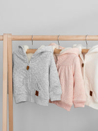 Drei Baby-Kapuzenjacken aus Baumwolle in Grau, Rosa und Weiß auf Holzständer.