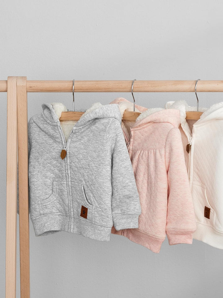 Drei Baby-Kapuzenjacken aus Baumwolle in Grau, Rosa und Weiß auf Holzständer.