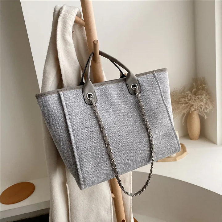 Graue Damenhandtasche aus Stoff mit Kettenriemen, elegant und modern, auf Holzständer.