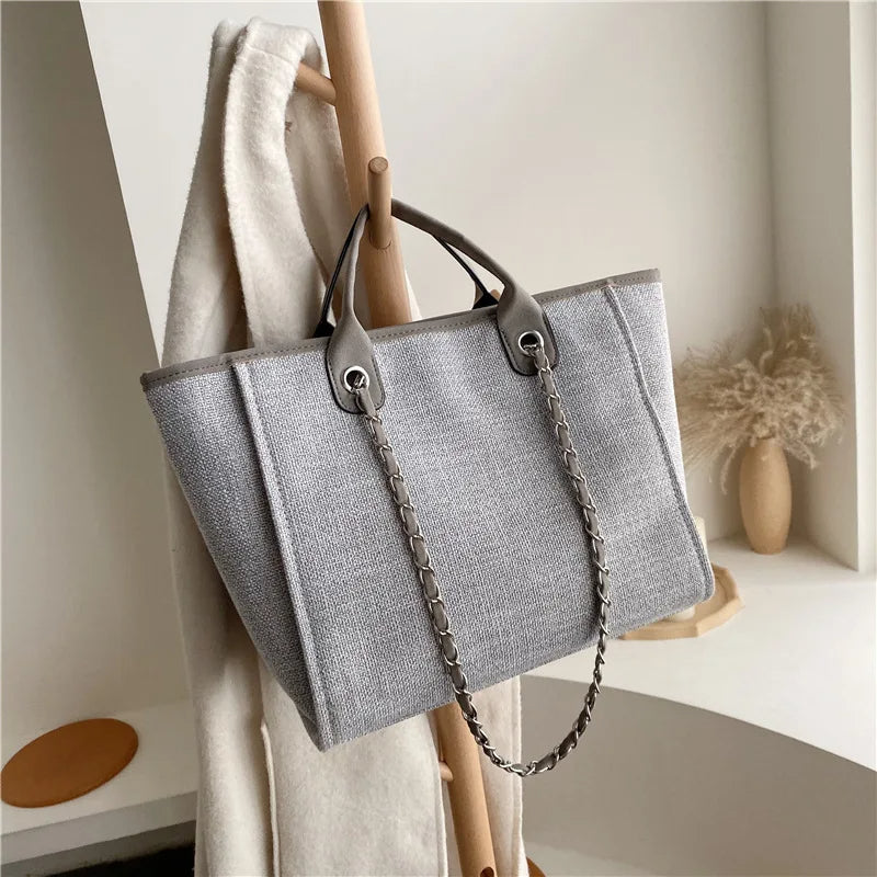 Graue Damenhandtasche aus Stoff mit Kettenriemen, elegant und modern, auf Holzständer.