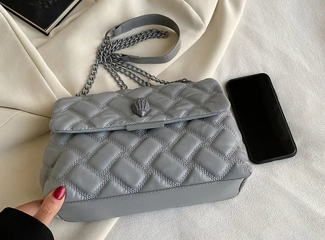 Graue gesteppte Handtasche aus Leder mit Kettengurt neben Smartphone auf weißem Tuch.