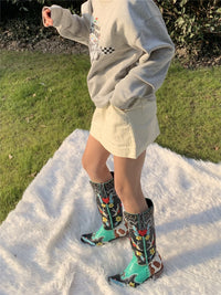 Frau in beigem Rock und grauem Pullover trägt bunte Cowboy-Stiefel auf weißem Teppich im Freien.