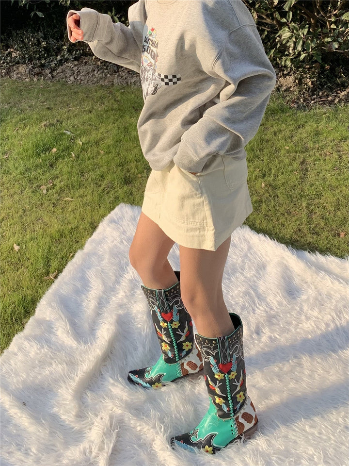 Frau in beigem Rock und grauem Pullover trägt bunte Cowboy-Stiefel auf weißem Teppich im Freien.