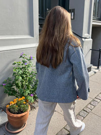 Frau in grauem Blazer und weißer Hose spaziert auf Bürgersteig neben Blumentopf.