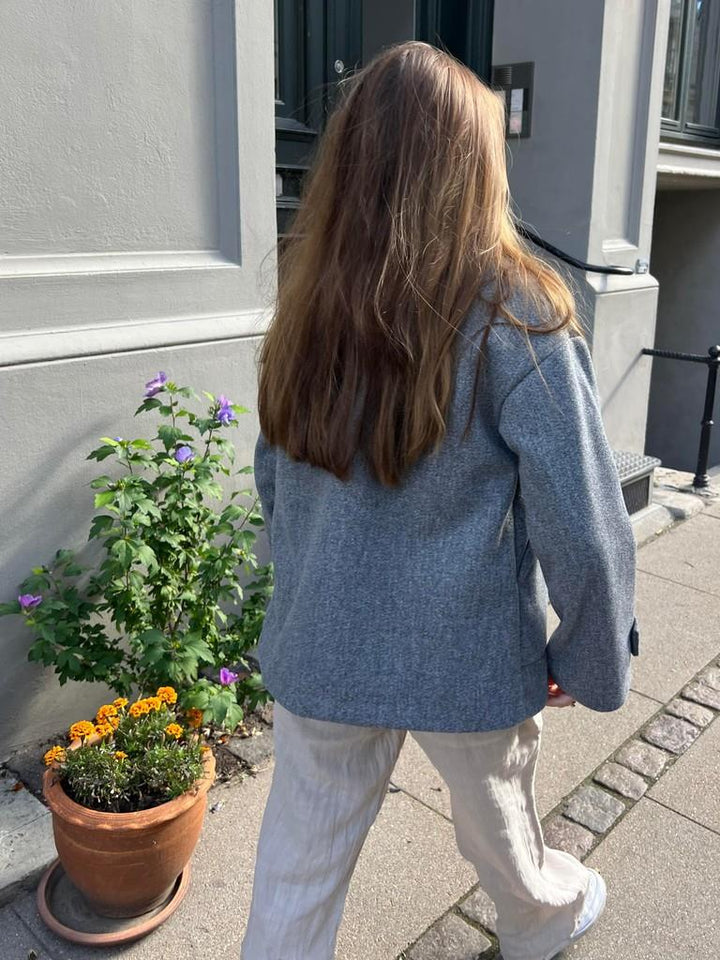 Frau in grauem Blazer und weißer Hose spaziert auf Bürgersteig neben Blumentopf.