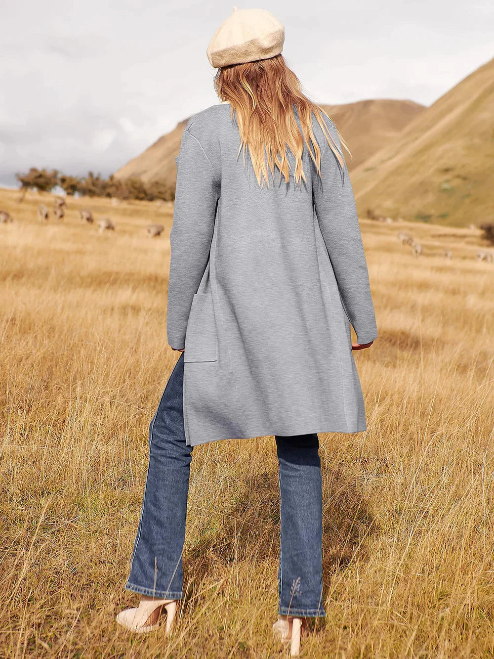 Frau in grauem Mantel und Jeans auf Wiese, beige Mütze, Herbstmode, Naturhintergrund.
