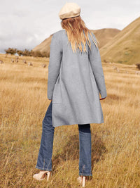 Frau in grauem Mantel und Jeans auf Wiese, beige Mütze, Herbstmode, Naturhintergrund.