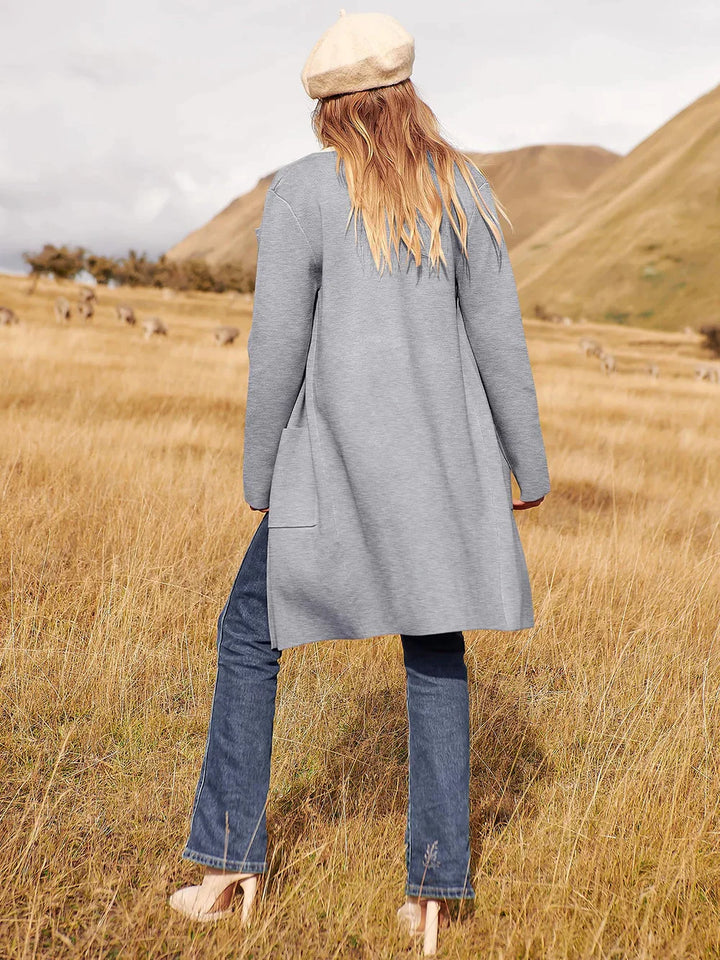 Frau in grauem Mantel und Jeans auf Wiese, beige Mütze, Herbstmode, Naturhintergrund.