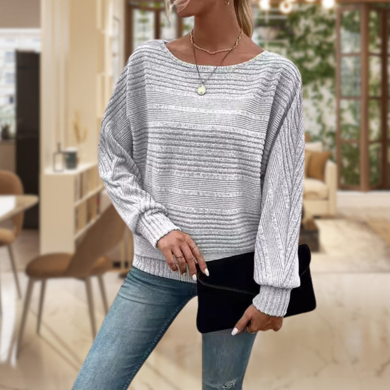 Frau in grauem Strickpullover und Jeans mit schwarzer Clutch in modernem Wohnzimmer.