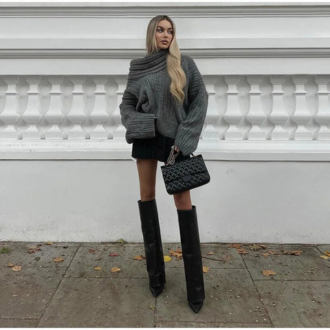 Frau in grauem Strickpullover mit einer Schulter, schwarze Stiefel, Tasche, Herbstmode.