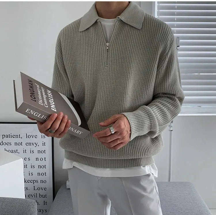 Mann in grauem Strickpullover mit Reißverschluss hält Buch, moderne Inneneinrichtung.