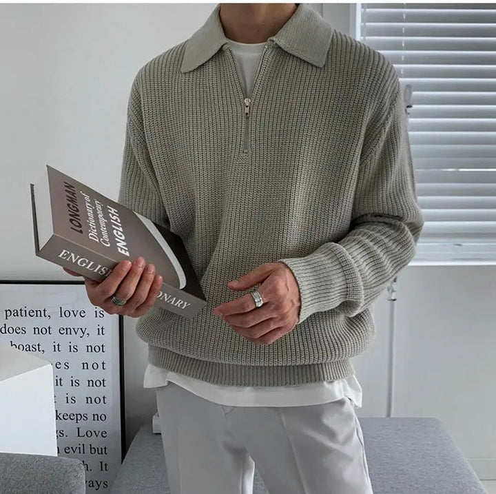 Mann in grauem Strickpullover mit Reißverschluss hält Buch, moderne Inneneinrichtung.