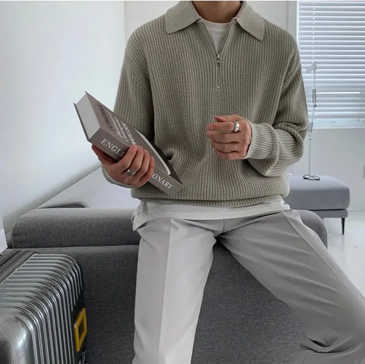 Mann in grauem Strickpullover mit Reißverschluss hält Buch, sitzt auf grauem Sofa.