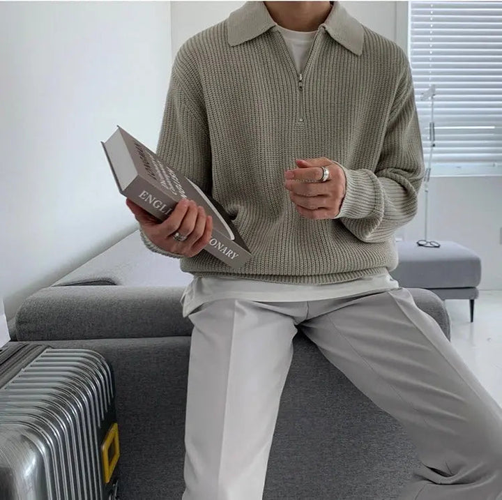 Mann in grauem Strickpullover mit Reißverschluss hält Buch, sitzt auf grauem Sofa.