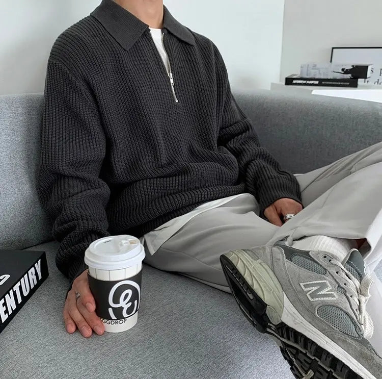 Mann in grauem Strickpullover mit Reißverschluss, hält Kaffeebecher, trägt graue Sneakers.