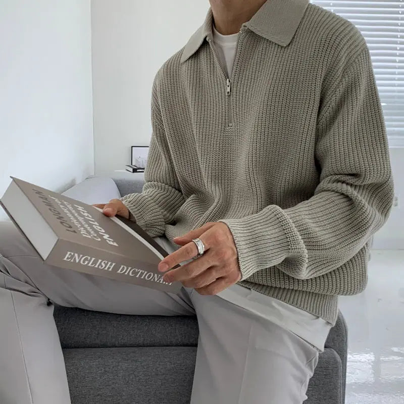 Mann in grauem Strickpullover hält englisches Wörterbuch, sitzt auf grauem Sofa.