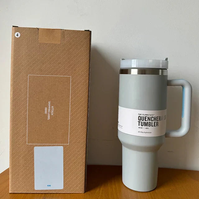 Grauer Stanley Quencher Tumbler 1,4L mit Griff, Edelstahl, neben brauner Verpackung.