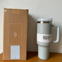 Grauer Stanley Quencher Tumbler 1,4L mit Griff, Edelstahl, neben brauner Verpackung.