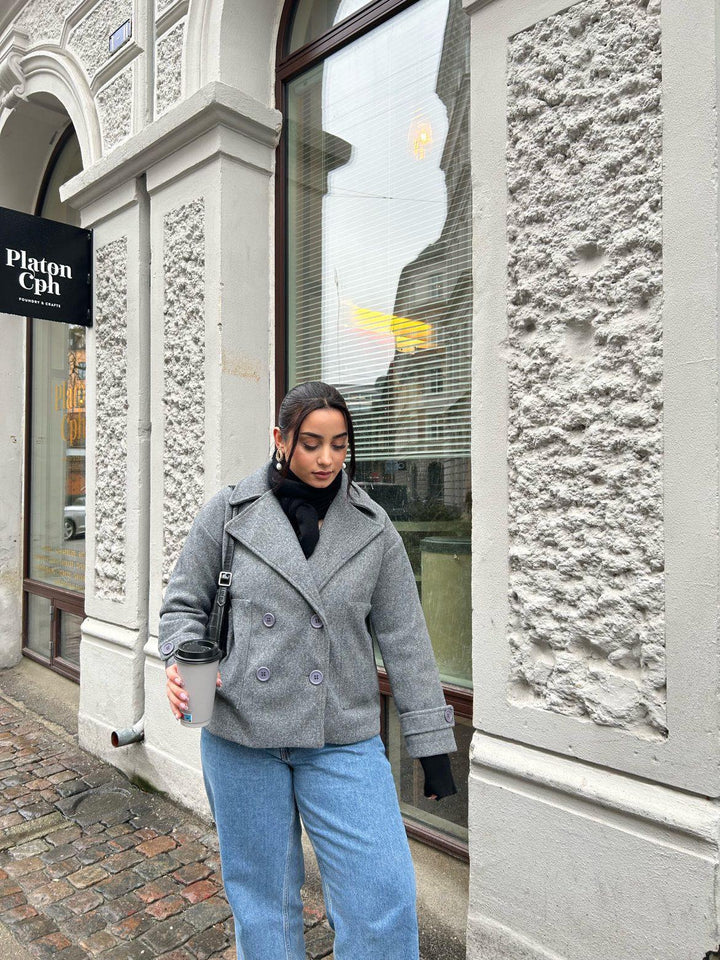 Frau in grauem Mantel mit Kaffeebecher vor Platon Cph Geschäft, modisch, urbaner Stil.