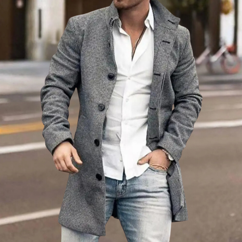 Mann in grauem Wollmantel, weißem Hemd und Jeans auf städtischer Straße.