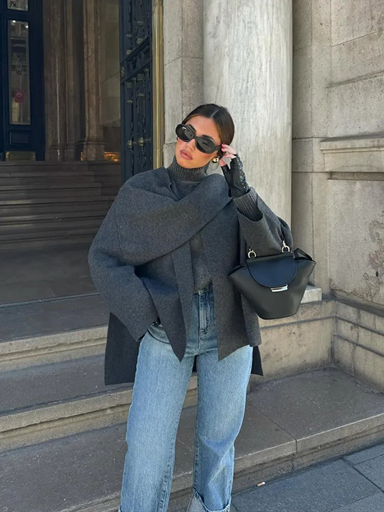 Frau in grauem Mantel und Jeans mit schwarzer Handtasche und Sonnenbrille vor Gebäude.