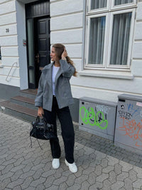 Frau in grauem Mantel und schwarzer Hose vor weißem Gebäude, modisch, urbaner Stil.