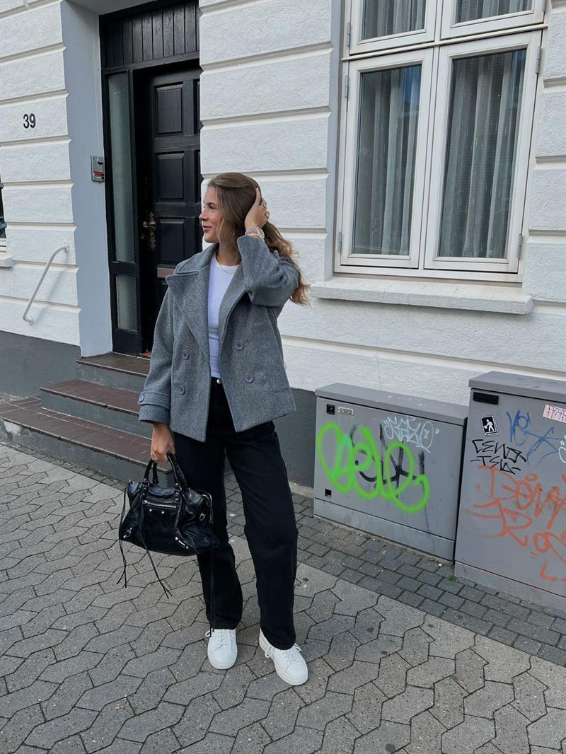 Frau in grauem Mantel und schwarzer Hose vor weißem Gebäude, modisch, urbaner Stil.