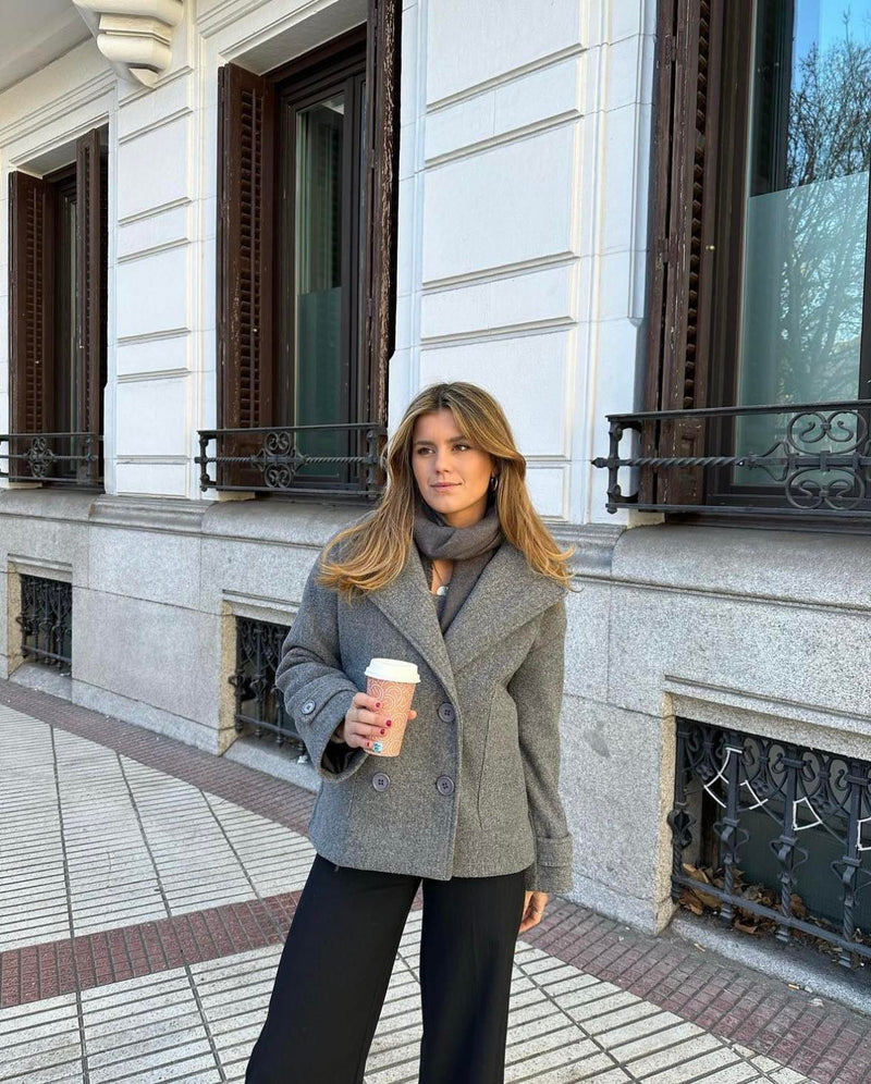 Frau in grauem Wollmantel mit Kaffeebecher, steht vor einem Gebäude, Herbstmode.