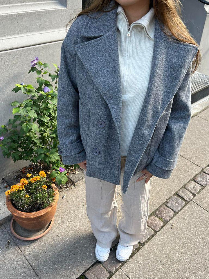 Frau in grauem Wollmantel, beiger Hose, weißen Schuhen, neben Blumentopf auf Gehweg.