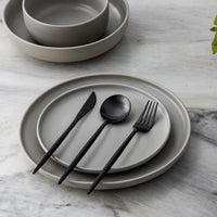 Graues Keramikgeschirr-Set mit schwarzem Besteck auf Marmortisch, modern und elegant.