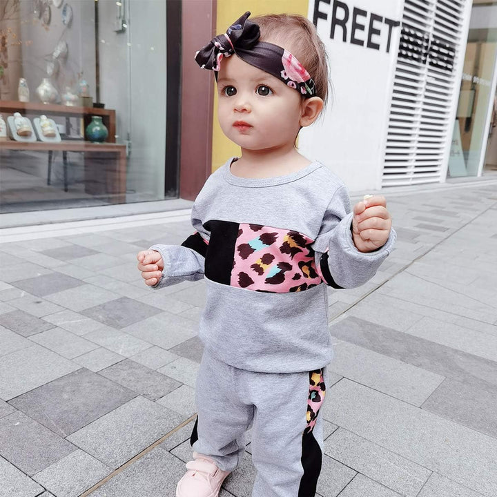 Kleinkind in grauem Jogginganzug mit Leopardenmuster, rosa Schleife, draußen auf Gehweg.