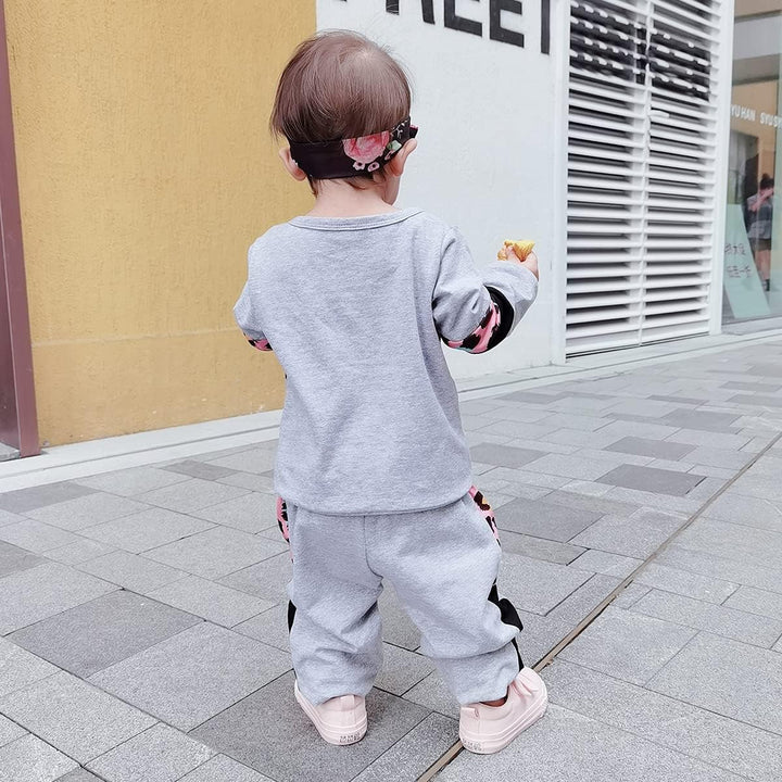 Kleinkind in grauem Jogginganzug mit rosa Details, draußen auf Gehweg, Rückansicht.