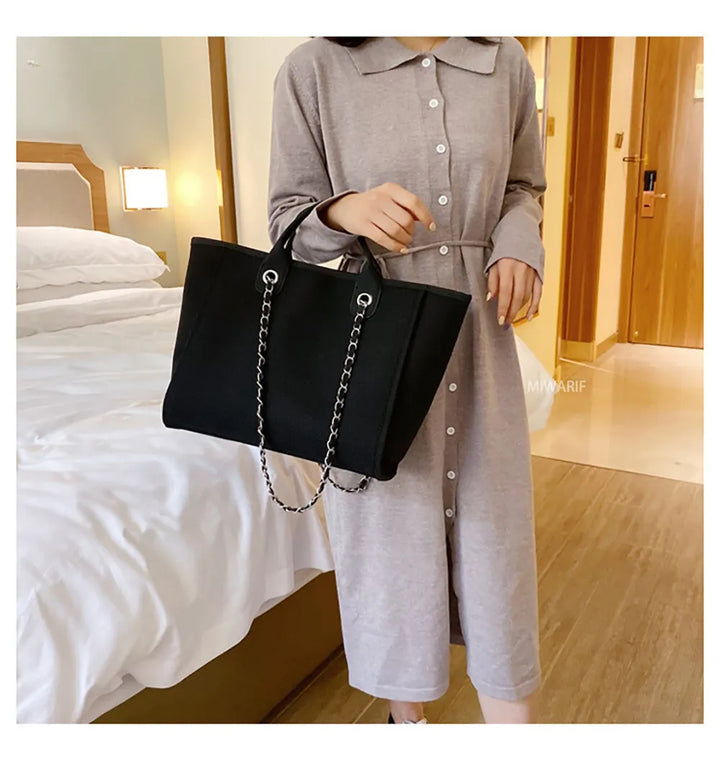 Frau in grauem Kleid mit schwarzer Handtasche aus Leder und Kette im Hotelzimmer.