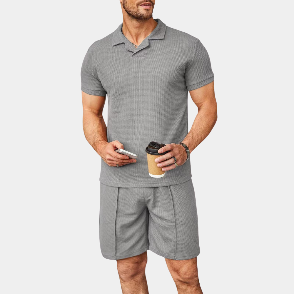 Mann in grauem Poloshirt und Shorts, hält Kaffeebecher und Smartphone.