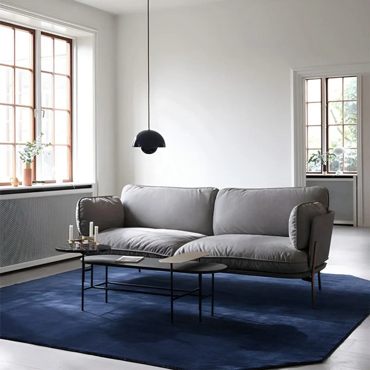 Graues Sofa im modernen Wohnzimmer, blauer Teppich, schwarze Hängelampe, große Fenster.