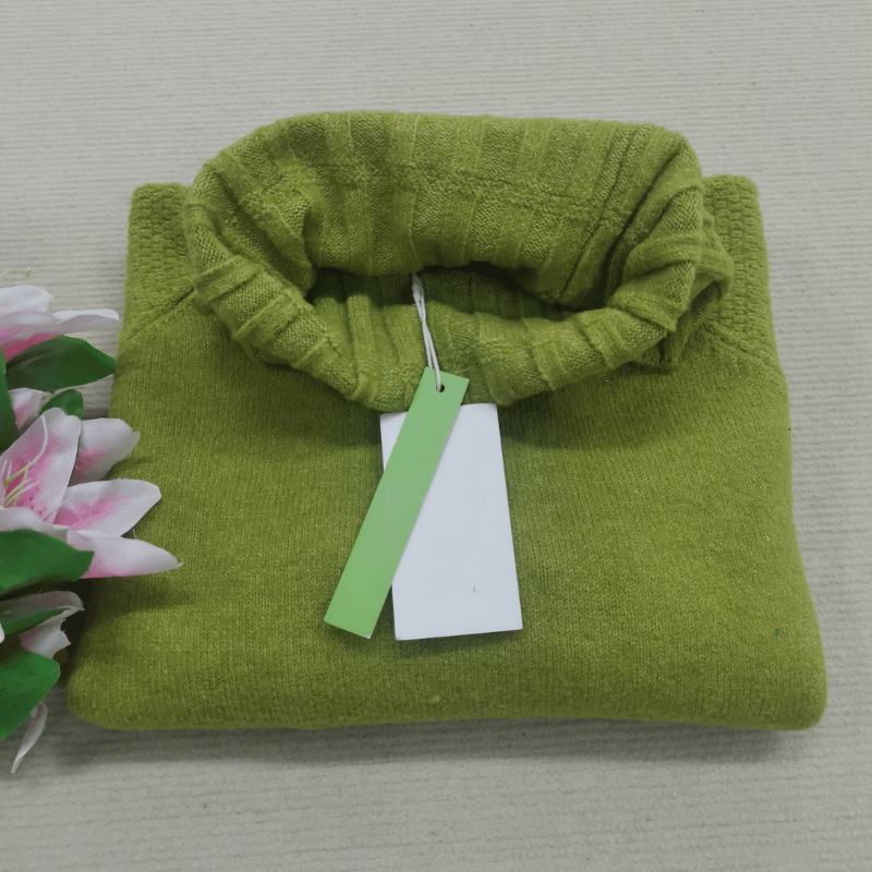 Grüner Rollkragenpullover aus Wolle, gefaltet, mit Etikett, neben rosa Blumen.