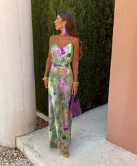 Frau im langen, floralen Sommerkleid mit lila Quasten-Ohrringen und Tasche, draußen stehend.