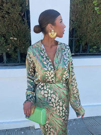 Frau in grünem gemustertem Kleid mit goldenen Ohrringen und grüner Clutch vor Hecke.