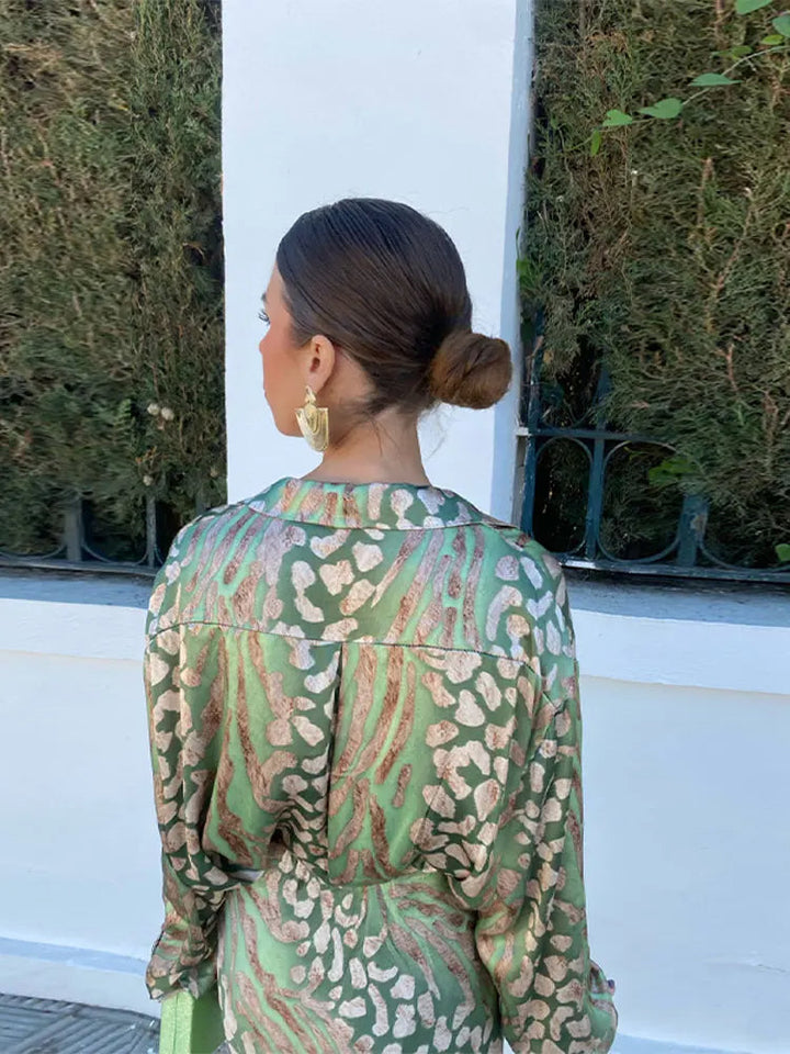 Frau in grünem Seidenkleid mit Leopardenmuster, goldene Ohrringe, eleganter Dutt, draußen.