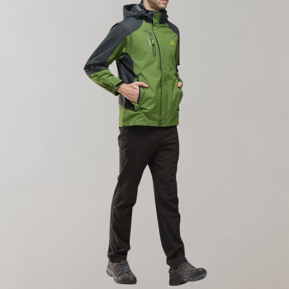 Mann in grüner Outdoor-Jacke mit Kapuze, schwarze Hose, Wanderschuhe, grauer Hintergrund.