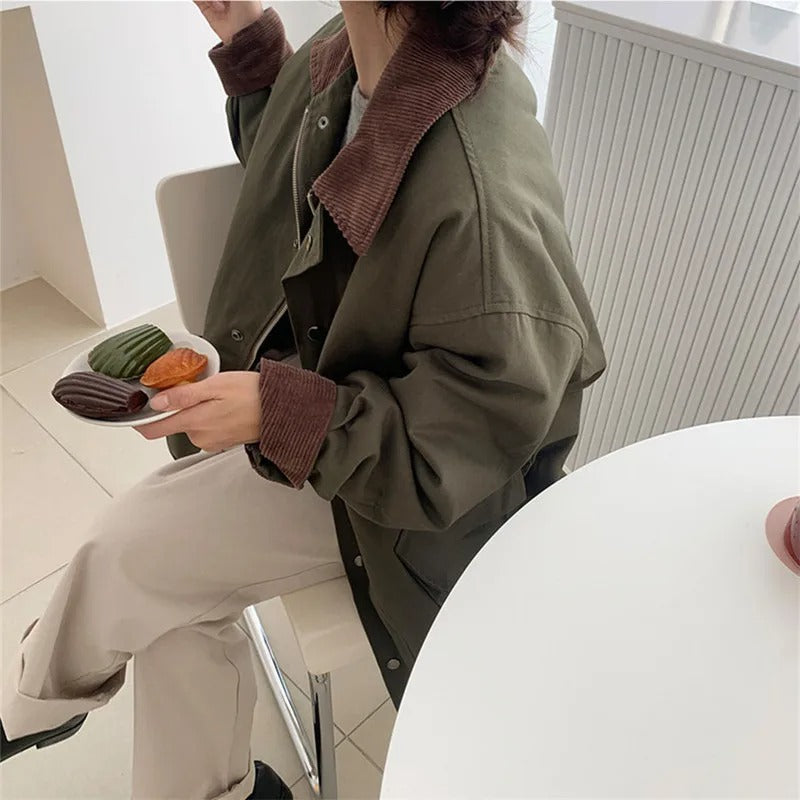 Person in olivgrüner Jacke mit braunem Kragen, hält Teller mit Gebäck, sitzt an weißem Tisch.