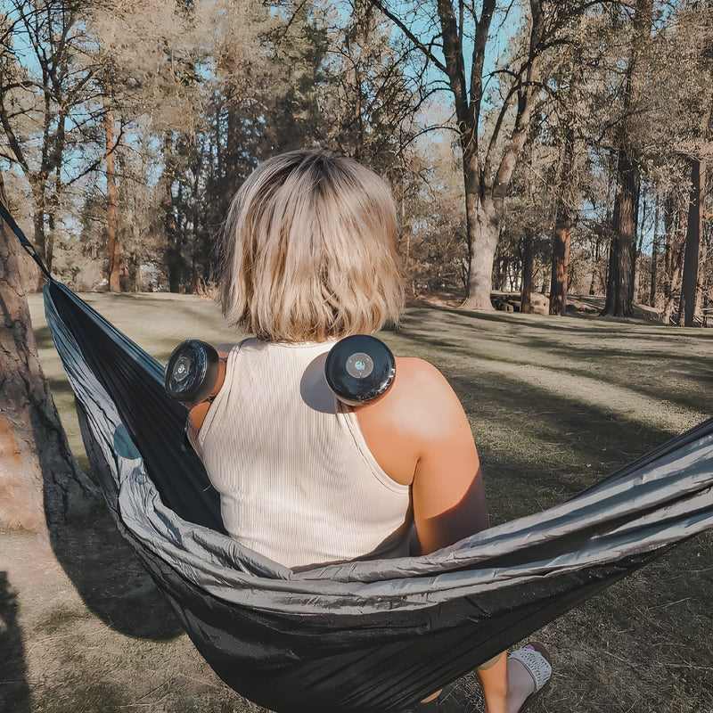Frau in Hängematte mit Massagegeräten auf Schultern im Wald, Entspannung, Outdoor, Freizeit.