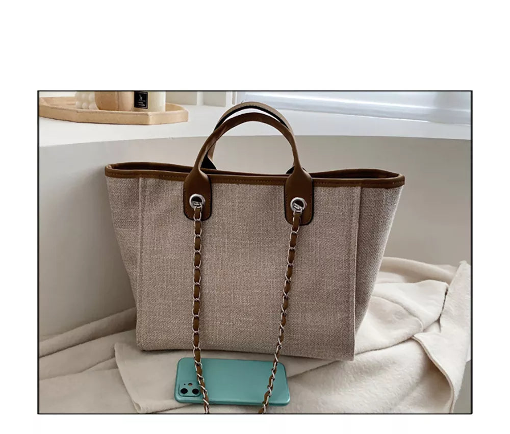 Beige Damenhandtasche aus Leinen mit braunen Ledergriffen und Kettendetails, neben Smartphone.