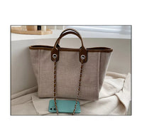 Beige Damenhandtasche aus Leinen mit braunen Ledergriffen und Kettendetails, neben Smartphone.